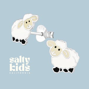 May include: A pair of silver sheep stud earrings with white enamel and black accents. The earrings are marked "925" for sterling silver. The earrings are from Salty Kids California.