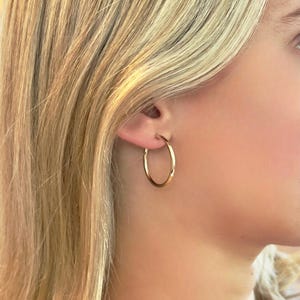 May include: A close-up of a person's ear wearing a gold hoop earring. The earring is a simple, circular design with a polished finish. The person has blonde hair and fair skin.