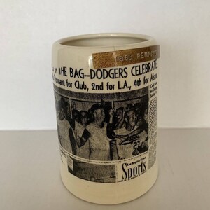 May include: A white ceramic mug with a black and white newspaper print design featuring a group of baseball players celebrating. The mug has a gold sticker with the text "1963 PENNANT" and "THE BAG--DODGERS CELEBRATE Pennant for Club, 2nd for L.A., 4th for Alston".