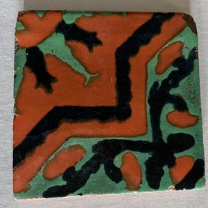May include: A square ceramic tile with a geometric design. The tile is decorated with black, orange, and green paint. The design features a series of abstract shapes and lines.