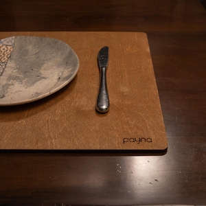 Puede incluir: Un mantel individual de madera marrón con un cuchillo de plata y un plato de cerámica blanco con un patrón gris y blanco. El mantel individual tiene la palabra "payma" impresa.