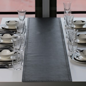 May include: A black leather table runner with a stitched border. The runner is on a white table with a set of glasses and dishes.