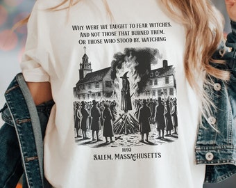 May include: A white t-shirt with a black and white illustration of a historical event in Salem, Massachusetts. The illustration depicts a person being burned at the stake with a crowd of people watching. The text on the shirt reads: "Why were we taught to fear witches, and not those that burned them, or those who stood by. Watching 1692 Salem, Massachusetts."