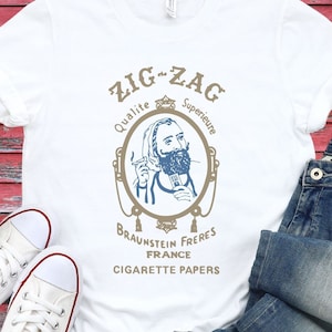 May include: White t-shirt with a vintage-style illustration of a man smoking a cigarette. The text on the shirt reads "Zig-Zag Qualite Superieure Braunstein Freres France Cigarette Papers".
