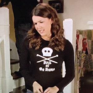 May include: A black long-sleeved shirt with a white skull and crossbones design. The shirt has the text "Knit or Go Home" printed below the skull.