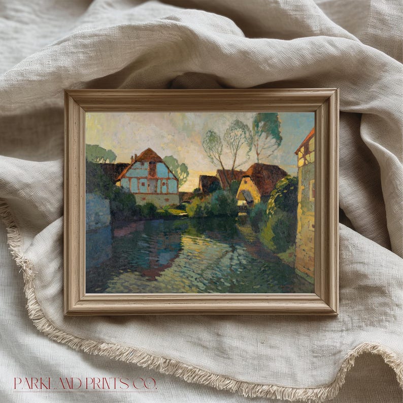 Framed European village print in a simple light wood frame, artistically arranged with flowing linen fabric in neutral tones.