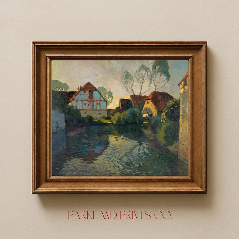 Classic village scene artwork displayed in an ornate gold-toned frame with decorative molding, mounted on a cream-colored wall.