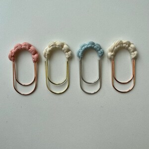 May include: Four decorative paper clips with a braided yarn design. The clips are in pink, white, light blue, and beige. Each clip has a double loop.