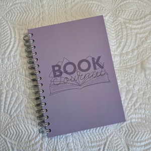 May include: A lavender spiral-bound journal with the words "BOOK Journal" printed on the cover. The text is above an illustration of an open book and mountains. The journal is lying on a white, textured surface.