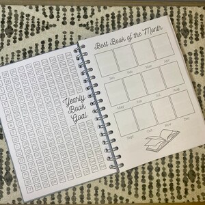 May include: A gray and white book with a spiral binding. The cover has the text "Best Book of the Month" and a grid of 12 boxes representing each month of the year. The left page has a numbered grid for tracking books read throughout the year. The text "Yearly Book Goal" is printed on the page.