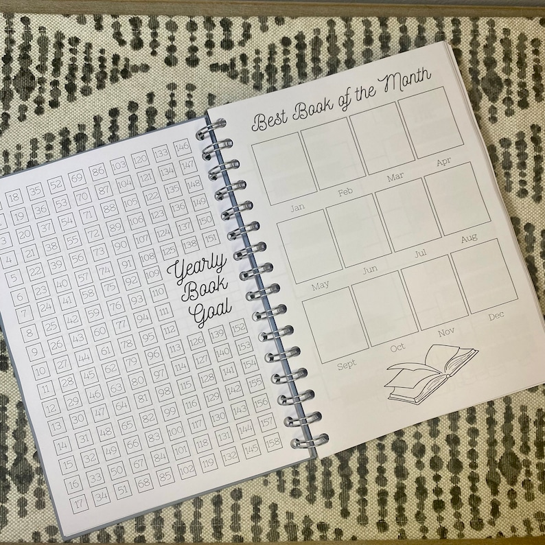 May include: A gray and white book with a spiral binding. The cover has the text "Best Book of the Month" and a grid of 12 boxes representing each month of the year. The left page has a numbered grid for tracking books read throughout the year. The text "Yearly Book Goal" is printed on the page.