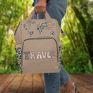 Puede incluir: Una mochila de lona beige con un diseño de mapa del mundo de estampado de leopardo y la palabra "TRAVEL" en letras rosas y blancas. La mochila tiene un bolsillo frontal con cremallera y correas ajustables.