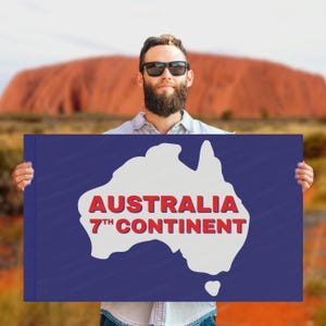 May include: A person holding a blue sign with a white outline of Australia and the words "AUSTRALIA 7TH CONTINENT" in red. The background features a large red rock formation and a clear sky.