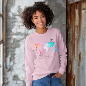 May include: A pink long-sleeved shirt with a watercolor world map design and the text "Adventurous Awaits" in a cursive font. The map is surrounded by a dotted line and a small airplane is flying above it.