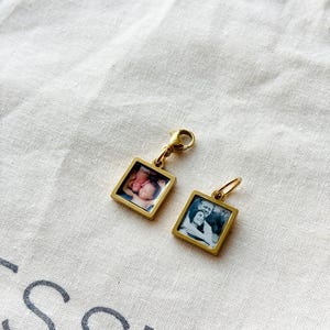 May include: Two gold-toned square pendants with small photographs inside. One pendant features a color photo of a baby, the other a black and white photo of a couple. Both have a small clasp for attaching to a chain or bracelet.