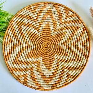 Pode incluir: Uma tapeçaria de parede redonda tecida com um padrão circular de cor bege e branco. A tapeçaria de parede tecida é feita de materiais naturais e tem um estilo rústico e boémio.