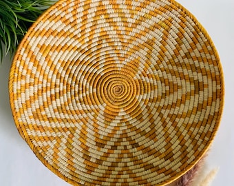 May include: A large, round woven basket with a yellow and white geometric pattern. The basket is made of natural materials and has a rustic, handcrafted look.
