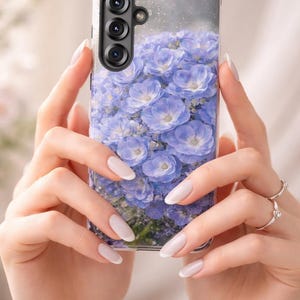 以下が含まれることがあります： 淡い青と紫のアジサイの花がプリントされた透明なスマートフォンケース。左上隅には3つのレンズを備えたカメラシステム用の長方形の切り欠きがあり、表面には繊細なグリッター仕上げが施されています。