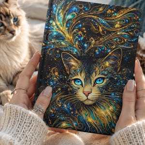 May include: A rectangular hardcover journal featuring a vibrant, intricate illustration of a cat with piercing blue eyes. The artwork displays swirling gold and teal patterns against a deep black background, creating a luminous, cosmic effect.