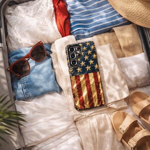 Puede incluir: Una funda para smartphone con un dise&ntilde;o brillante de la bandera estadounidense con estrellas doradas y rayas rojas y blancas, colocada dentro de una maleta con ropa doblada, un sombrero de paja, gafas de sol marrones y sandalias beige.