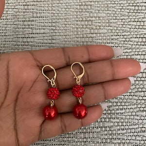 May include: A pair of red dangle earrings with gold hooks. The earrings feature a small red rhinestone ball and a larger red pearl.