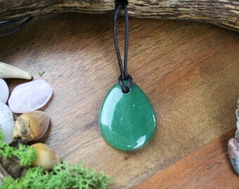 May include: A green aventurine gemstone pendant on a black leather cord. The pendant is teardrop shaped and is surrounded by natural elements like antlers, pine cones, and moss.