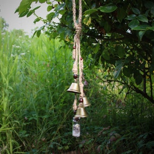 May include: A wind chime with multiple brass bells hanging from a rope with brown beads. The chime is hanging from a tree branch in a green grassy area.