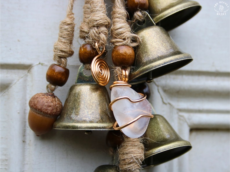 Pode incluir: Um m&oacute;bile de vento com v&aacute;rias sinos, contas de madeira castanhas e um cristal de quartzo transparente envolto em fio de cobre. Os sinos est&atilde;o presos a um cord&atilde;o castanho grosso.