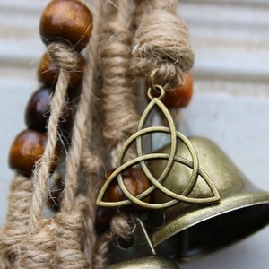 Pode incluir: Um m&oacute;bile marrom e dourado com contas de madeira, corda de juta e um amuleto de n&oacute; celta. O amuleto est&aacute; preso a um sino.
