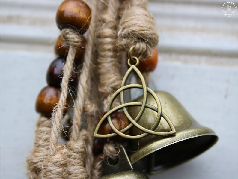 Pode incluir: Um m&oacute;bile marrom e dourado com contas de madeira, corda de juta e um amuleto de n&oacute; celta. O amuleto est&aacute; preso a um sino.