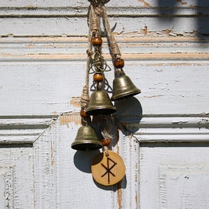 Könnte beinhalten: Ein Holzanhänger mit einem geschnitzten Symbol hängt an einer Glockenkette. Die Glocken sind golden und haben ein rustikales Aussehen. Der Anhänger ist braun und hat eine helle Holzmaserung. Der Hintergrund ist eine weiße Holzoberfläche.