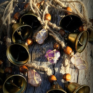 May include: A collection of brass bells with amethyst crystals hanging from twine and wooden beads. The bells are arranged on a rustic wooden surface.