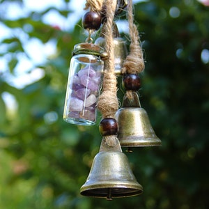 May include: A wind chime with three brass bells and a glass vial filled with amethyst crystals. The chime is hung from a length of twine with wooden beads.