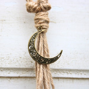 Pode incluir: Uma tape&ccedil;aria de parede de macram&eacute; de corda de juta marrom com um amuleto de lua crescente dourado.