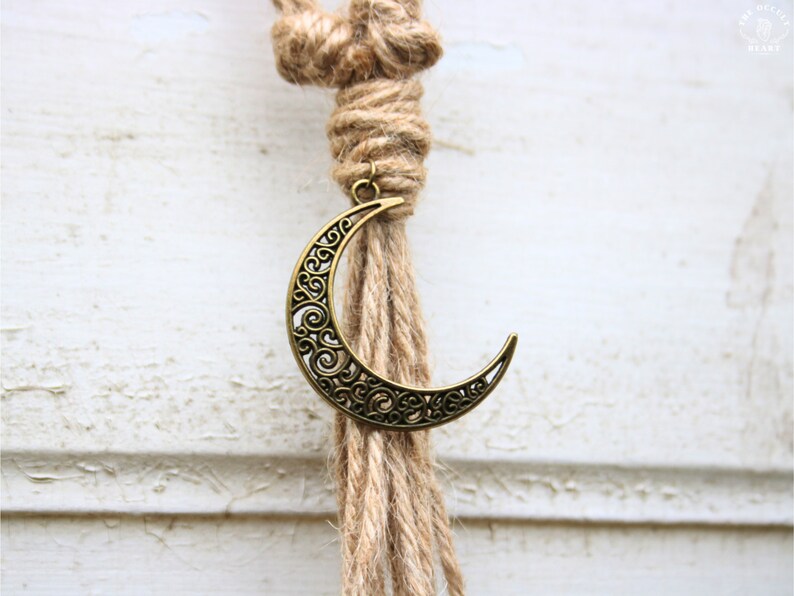 Pode incluir: Uma tape&ccedil;aria de parede de macram&eacute; de corda de juta marrom com um amuleto de lua crescente dourado.
