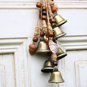 Pode incluir: Uma corda de sinos com um amuleto de cristal pendura em uma porta branca. Os sinos s&atilde;o de lat&atilde;o e t&ecirc;m um visual r&uacute;stico e vintage. A corda &eacute; de barbante e tem contas de madeira e bolotas.