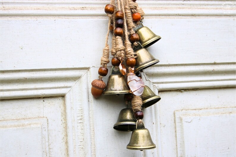 Pode incluir: Uma corda de sinos com um amuleto de cristal pendura em uma porta branca. Os sinos s&atilde;o de lat&atilde;o e t&ecirc;m um visual r&uacute;stico e vintage. A corda &eacute; de barbante e tem contas de madeira e bolotas.