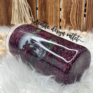 May include: A purple glitter tumbler with a skull and roses design. The tumbler is on a white fluffy surface. The text "creative design outlet" is written in black cursive on the tumbler.