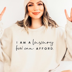Puede incluir: Una sudadera blanca con el texto "I AM A LUXURY FEW CAN AFFORD." impreso en negro.