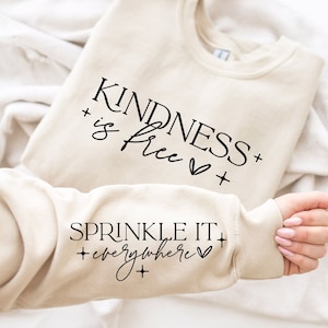 以下が含まれることがあります： ベージュのトレーナーに「KINDNESS is free」という文字とハートが前面に、袖には「SPRINKLE IT everywhere」という文字とハートがプリントされています。