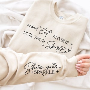 Op de afbeelding: Een beige sweatshirt met de tekst "never let anyone dull your sparkle" op de voorkant en "share your sparkle" op de mouw.