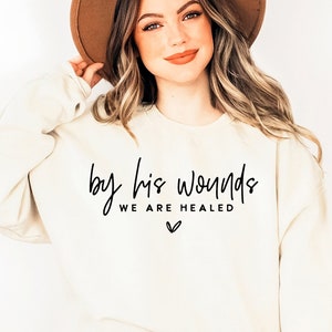 May include: A white sweatshirt with the text "by his wounds we are healed" in black lettering. The sweatshirt has a small heart symbol below the text.
