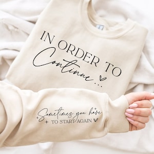 以下が含まれることがあります： ベージュのトレーナー。前面に「IN ORDER TO CONTINUE...」と、袖に「Sometimes you have to START AGAIN」の文字が書かれている。