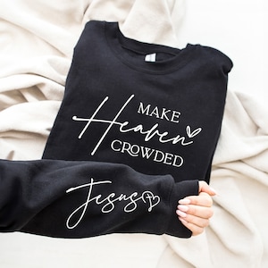 Puede incluir: Sudadera negra con texto blanco que dice "Make Heaven Crowded" y un s&iacute;mbolo de coraz&oacute;n. La manga de la sudadera tiene texto blanco que dice "Jesus" y un s&iacute;mbolo de coraz&oacute;n.