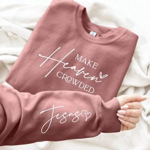 Puede incluir: Una sudadera de color rosa polvoriento con el texto "Make Heaven Crowded" y un coraz&oacute;n en la parte delantera y el texto "Jesus" y una cruz con un coraz&oacute;n en la manga.