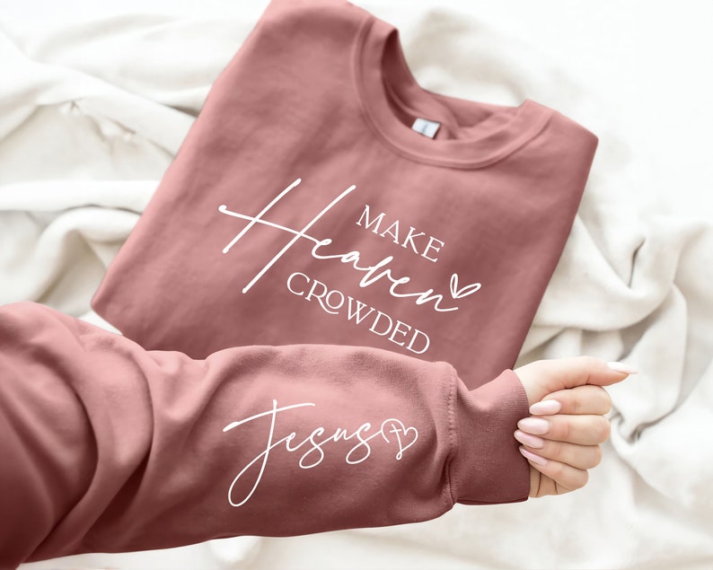 Puede incluir: Una sudadera de color rosa polvoriento con el texto "Make Heaven Crowded" y un coraz&oacute;n en la parte delantera y el texto "Jesus" y una cruz con un coraz&oacute;n en la manga.