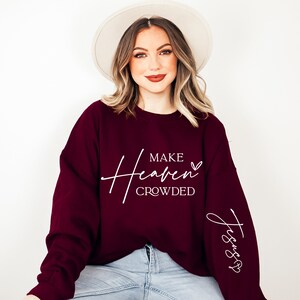 Puede incluir: Una sudadera color burdeos con el texto "Make Heaven Crowded" y "Jesus" en la manga. La sudadera la lleva puesta una persona con el pelo largo rubio y un sombrero blanco.