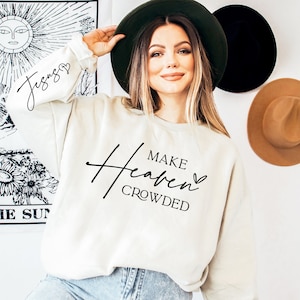 Puede incluir: Una sudadera blanca con el texto "Make Heaven Crowded" en escritura negra con un peque&ntilde;o s&iacute;mbolo de coraz&oacute;n. La modelo lleva la sudadera y un sombrero de ala ancha.