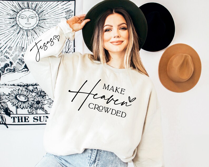 Puede incluir: Una sudadera blanca con el texto "Make Heaven Crowded" en escritura negra con un peque&ntilde;o s&iacute;mbolo de coraz&oacute;n. La modelo lleva la sudadera y un sombrero de ala ancha.