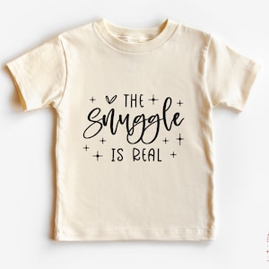 May include: A cream colored t-shirt with black text that reads "The + ♥ + Snuggle +  + Is Real + "